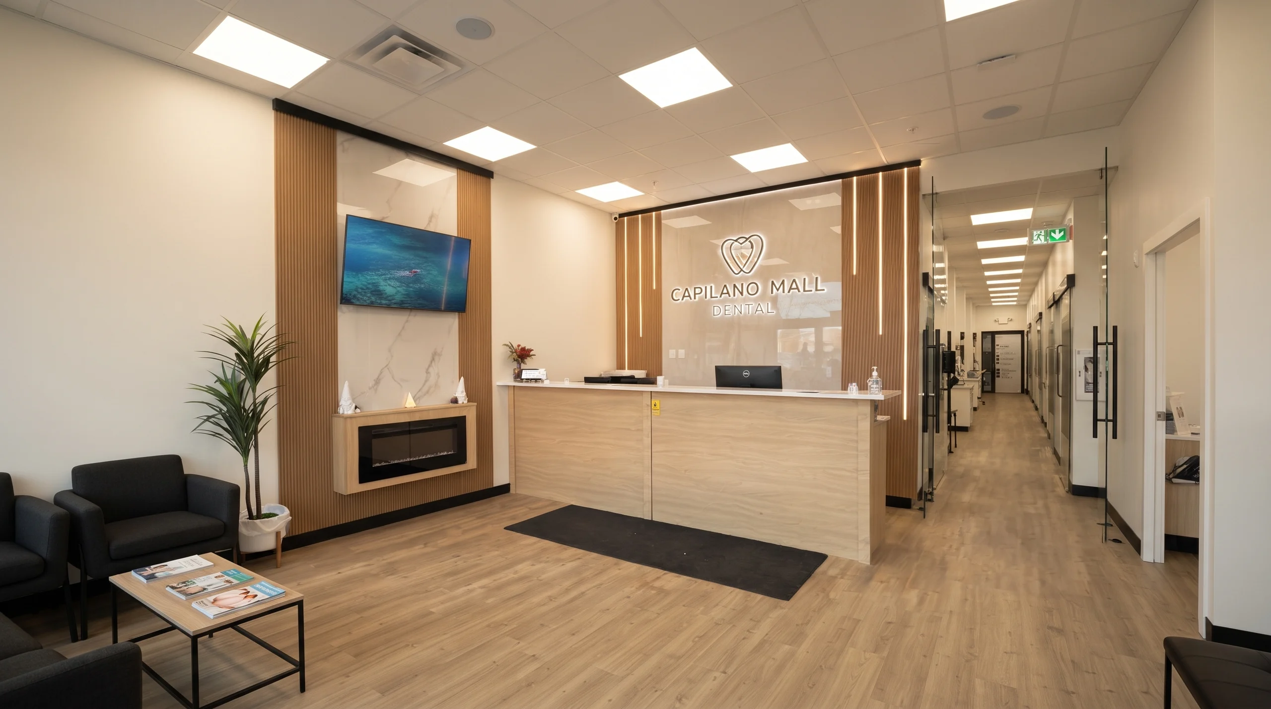 Reception area at Capilano Mall Dental in Edmonton, with the illuminated clinic sign, fireplace feature wall and treatment-room hallway beyond