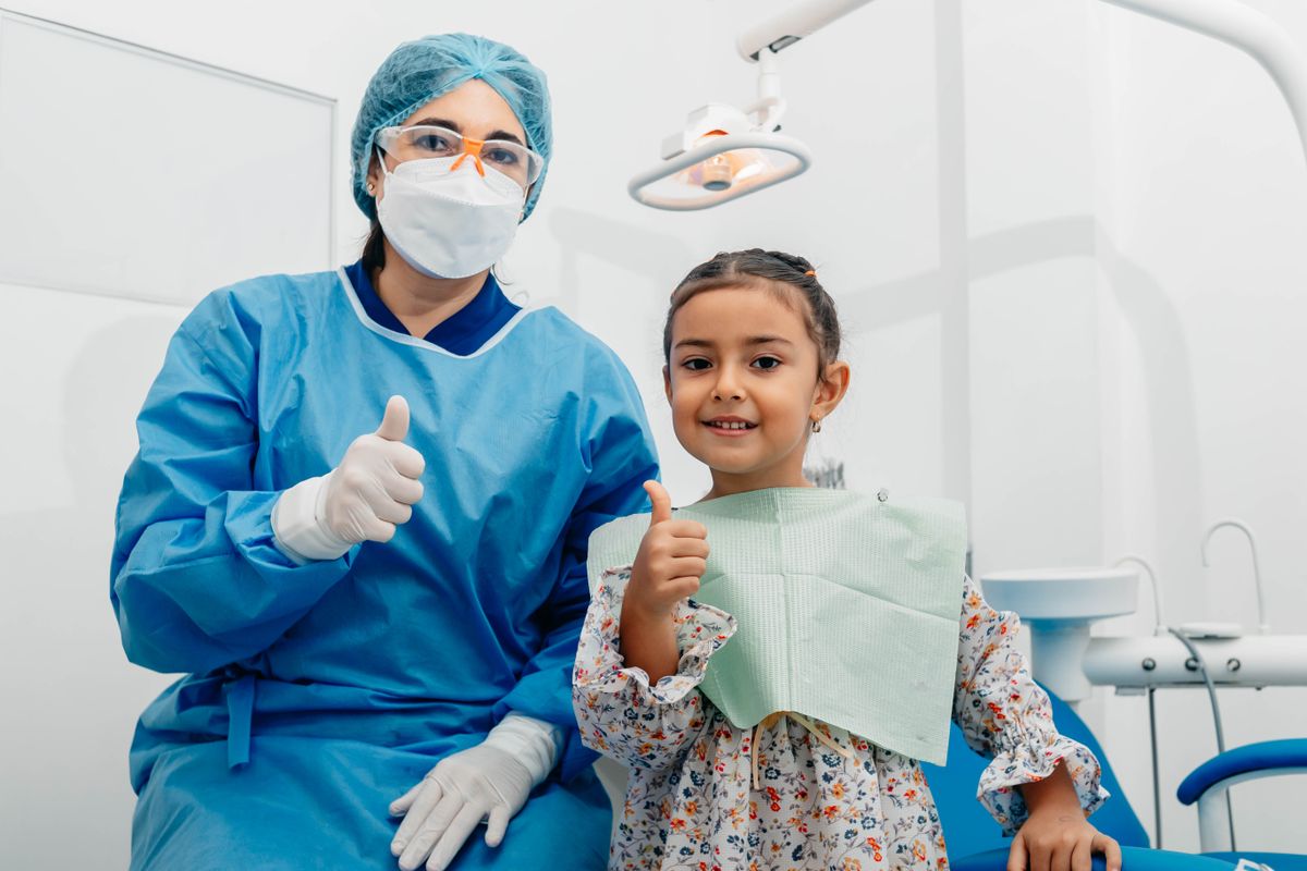 Child giving a thumbs-up in a dental chair beside a dental professional